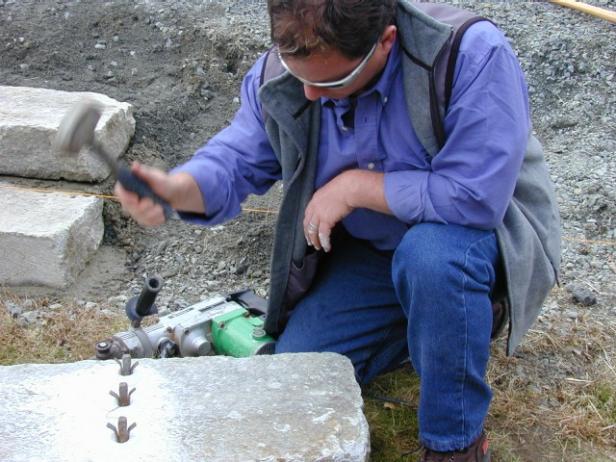To cut granite, first mark a straight and square line across the width of the stone. With a hammer gun, drill 3-inch-deep holes every 4 inches along the cutting line. Insert two feathers and a wedge into each hole. Pound the wedges with a stone hammer until the stone splits.