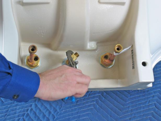 With the hardware mounted on the topside of the sink, mount the supply lines underneath. Screw the supply tee to the bottom of the spout, and then tighten it up with a pair of slip joint pliers.