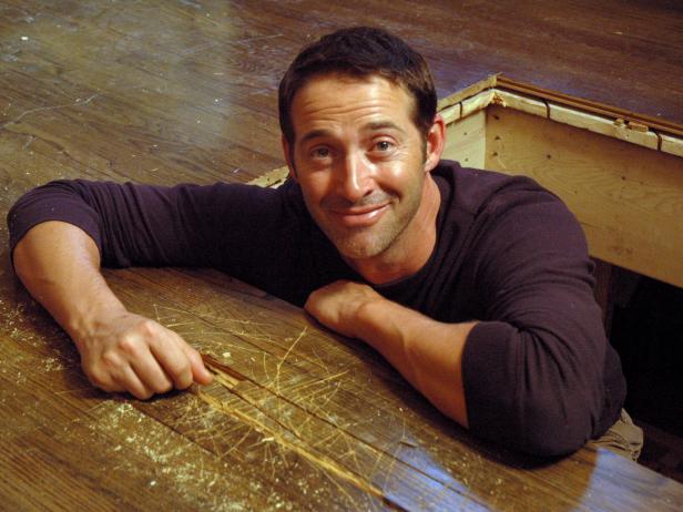 Josh Temple, host of Disaster House, crawls out of the hole in the hardwood floor to smile at the camera.