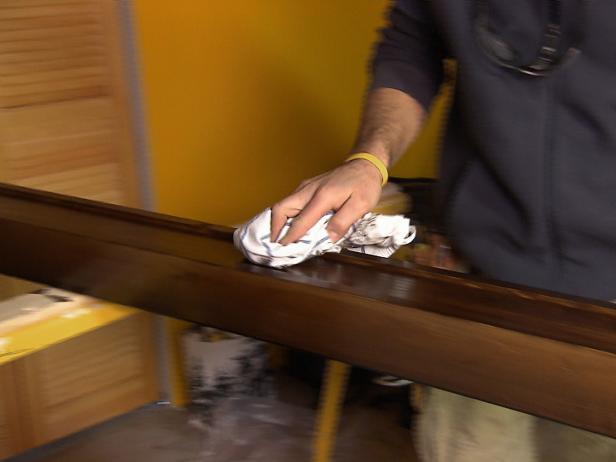 Man uses a rag to remove the excess stain that has been added to the long wooden piece.