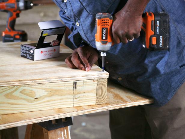 Closeup of man using a drill to attach plywood to base frame of doghouse construction project.