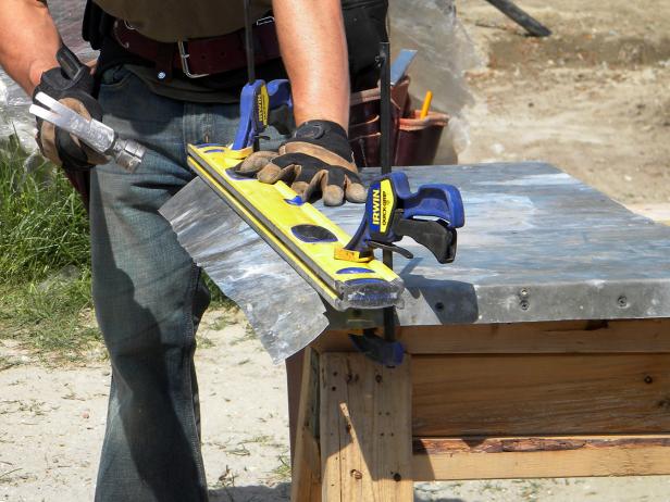 Lay the metal sheet on top of the plywood surface and hammer the metal sheet flat. Using wood clamps and a 2X4 (or level), temporarily secure the metal in place on the edge and bend over sides carefully with light hammer taps. Cut the metal at the corners to allow for a complete, flat wrap. The front metal wrap will fold over the side and attach underneath, out of sight. Nail the sides and front metal wrap in place.