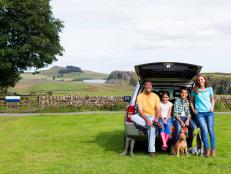 A family are posing for a picture in the countryside at the back of their car. Mother and dad are posing with their two children and their dog. A scenic landscape can be seen in the background.