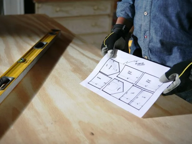 Man in bluejean shirt holds doghouse construction blueprints while standing next to wood table and level.