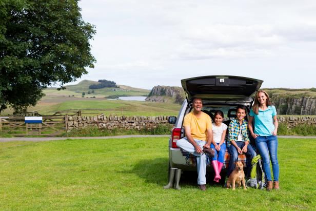 A family are posing for a picture in the countryside at the back of their car. Mother and dad are posing with their two children and their dog. A scenic landscape can be seen in the background.