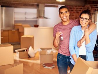 a young couple unpack their belongings as they settle into their new loft apartment . They are hugging and looking to camera smiling holding their keys aloft .