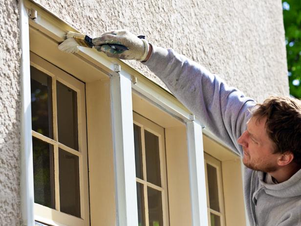 A professional house painter, small construction business entrepreneur, home owner or handyman works on exterior wooden surface, painting house window trim for repair and home improvement. The working man wears a painting cap, protective work gloves, and hooded sweatshirt, carefully brushing the paintbrush to apply latex paint for fall maintenance.